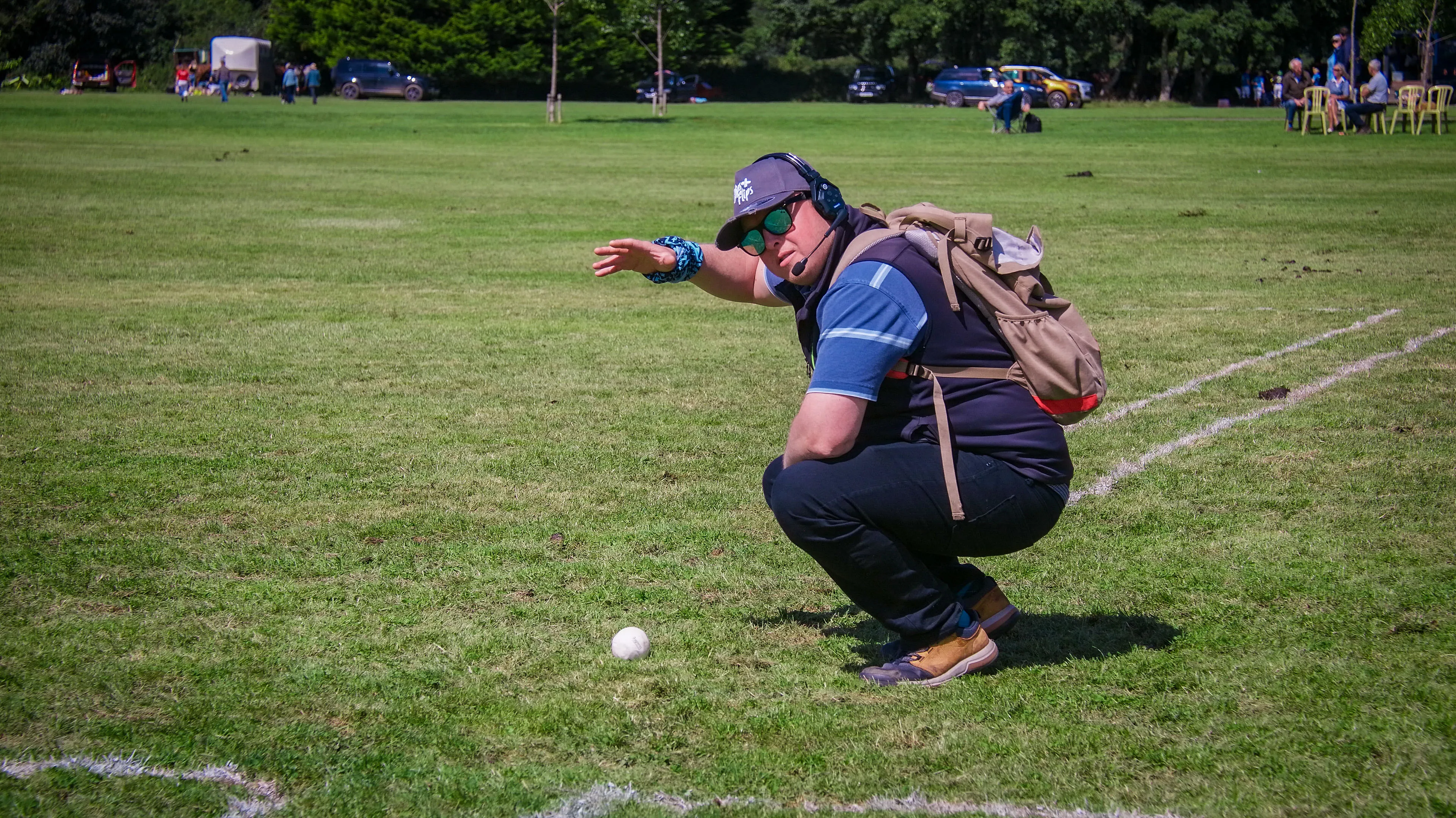 A person wearing sunglasses and headphones squats on a grassy field, pointing at a white ball. They appear engaged, with a relaxed outdoor vibe.